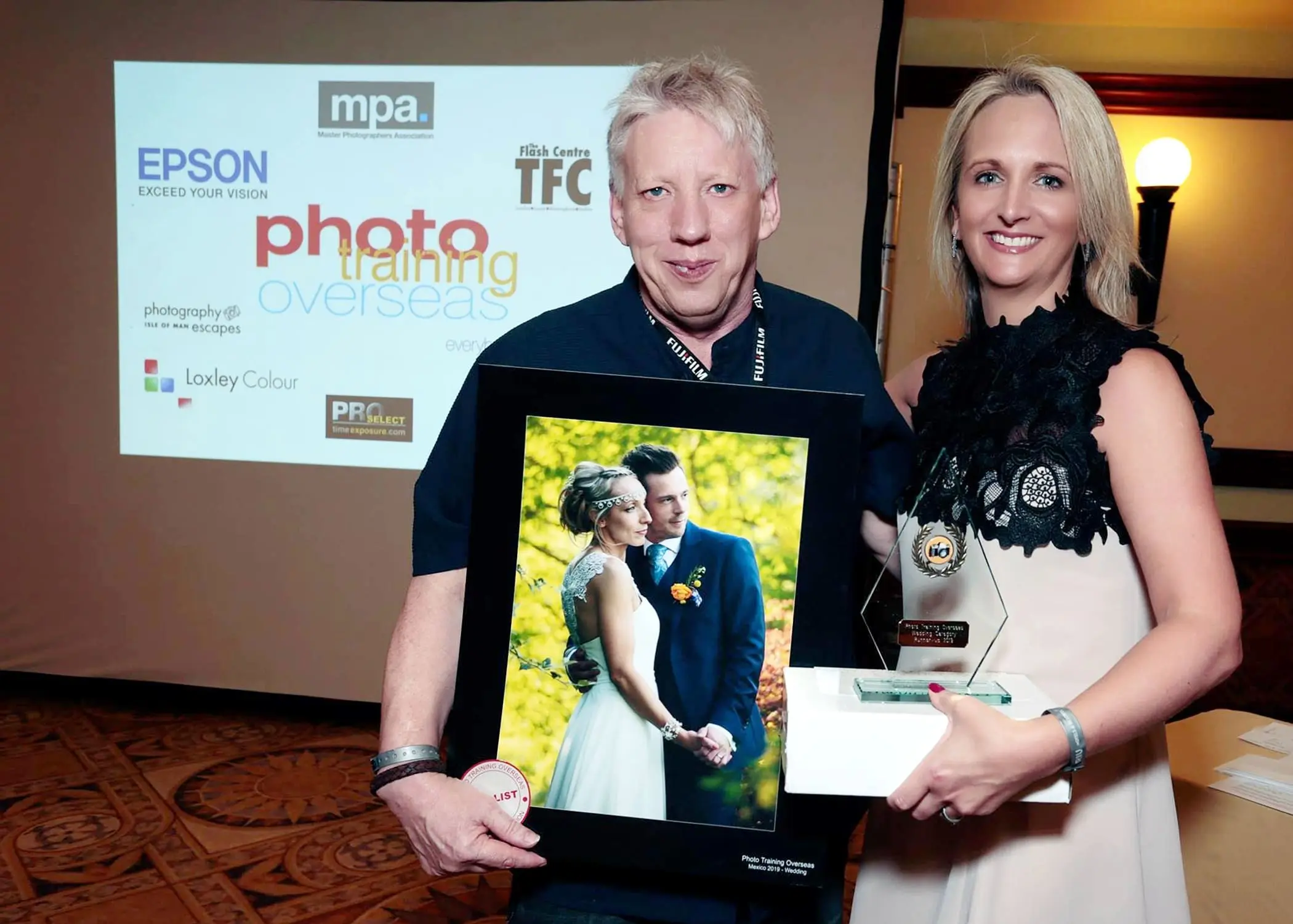 Two people holding photography award and photo. Event backdrop.